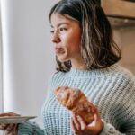 Attentive Asian female standing near curtains with delicious croissant in kitchen and looking away