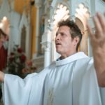 Priest with raised hands leading a Christian worship service inside a decorated church.