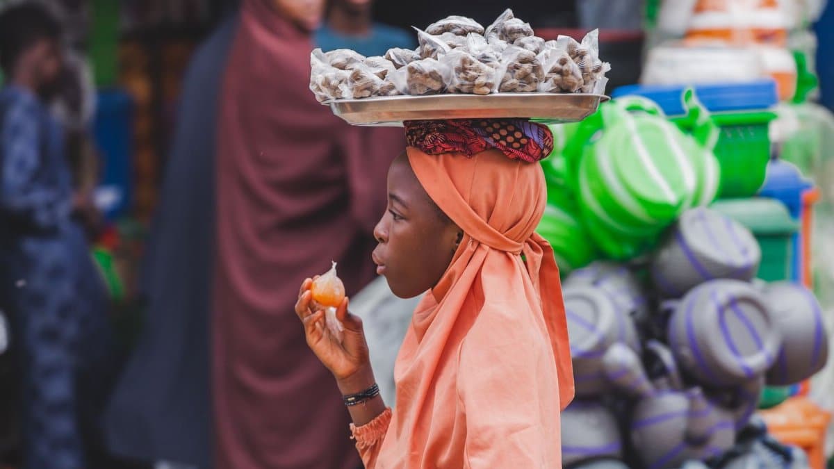 A young street vendor balances goods on her head in a vibrant market scene.