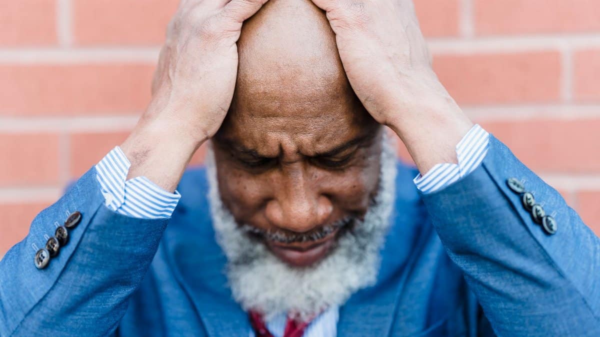 Unhappy bald African American male worker with gray beard in classy wear standing near building with hands on head on street