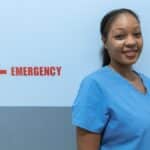 Happy healthcare worker in scrubs standing near emergency room sign.