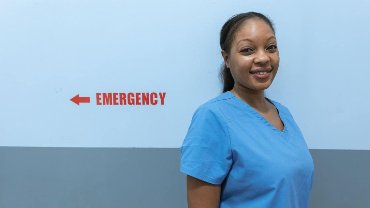 Happy healthcare worker in scrubs standing near emergency room sign.