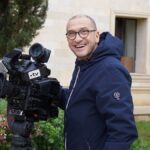 Smiling cameraman with a large TV camera in an outdoor setting near a building.