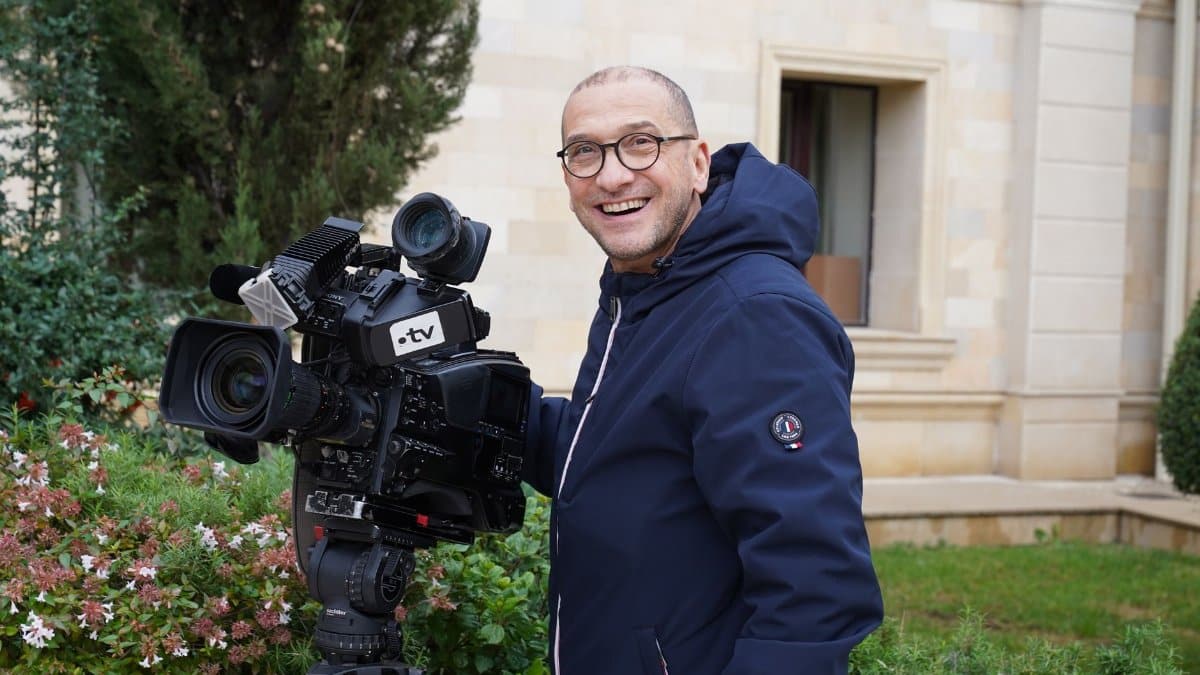 Smiling cameraman with a large TV camera in an outdoor setting near a building.