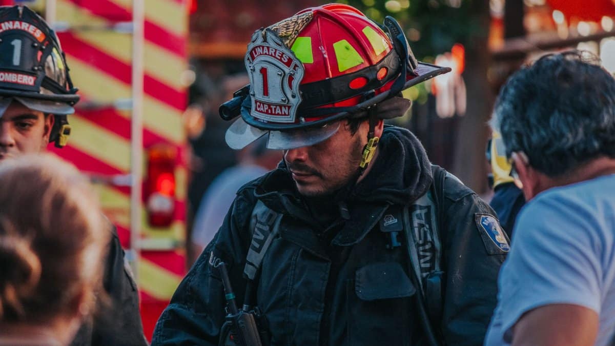 Firefighter attending an emergency on city streets, wearing uniform and helmet, with emergency vehicles in background.