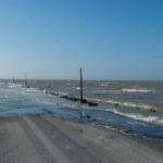 A flooded coastal road surrounded by the sea and horizon under a clear blue sky.