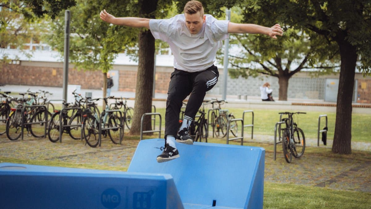 A young man jumping over blue parkour equipment in a park with bikes in the background.