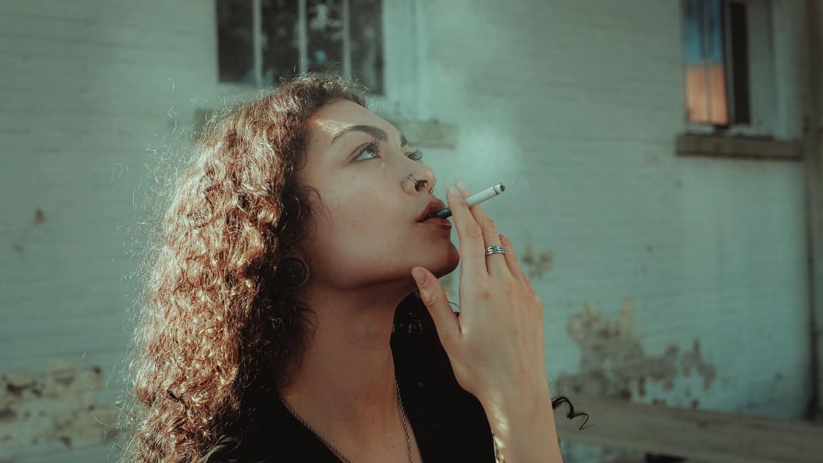 Young woman with cigarette, looking pensive while smoking outdoors.