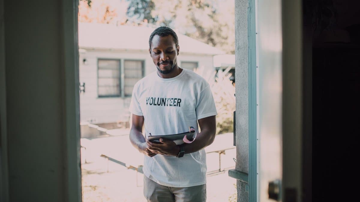 A man wearing a volunteer t-shirt stands by a doorway holding a tablet, engaged in a community event.