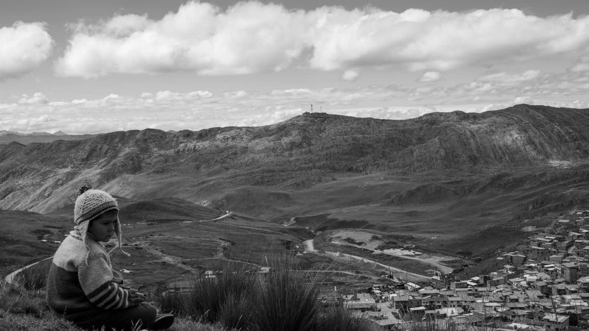 Child in traditional clothing sitting on a hill overlooking Cerro De Pasco's dramatic mountainous landscape.
