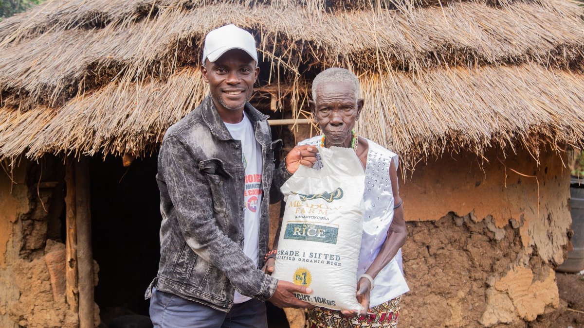 Two men displaying rice donation in front of a thatched hut in a rural village.