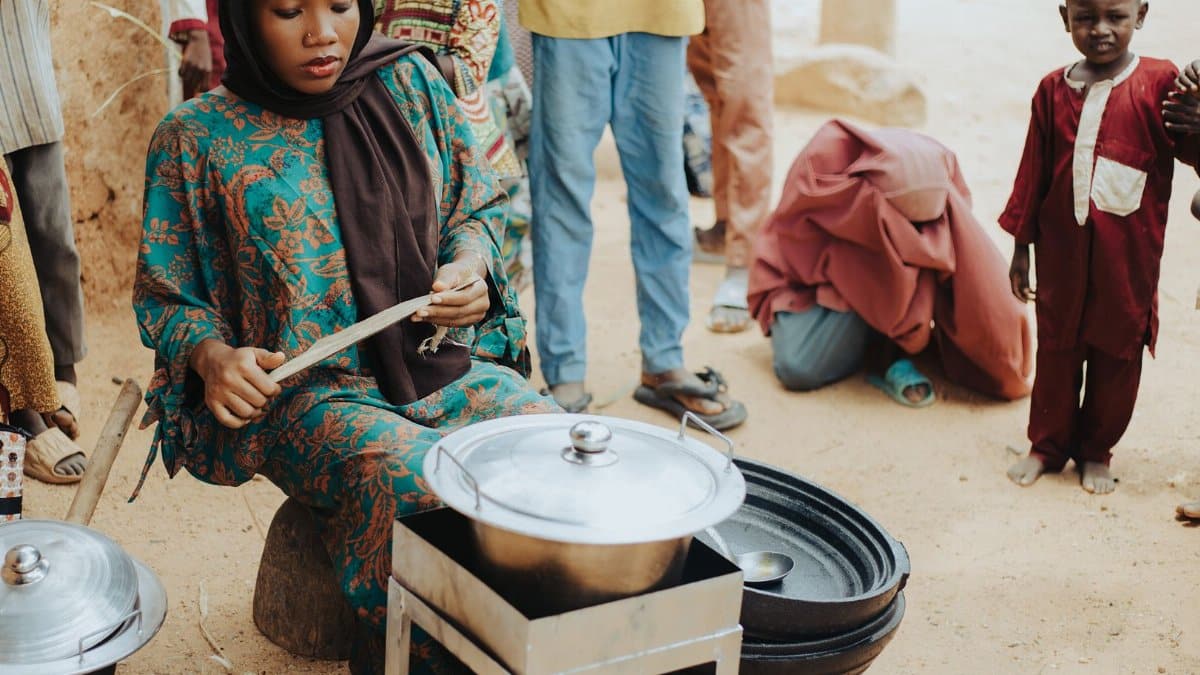 Young woman cooking outdoors in a community setting, surrounded by people. Diverse cultural atmosphere.