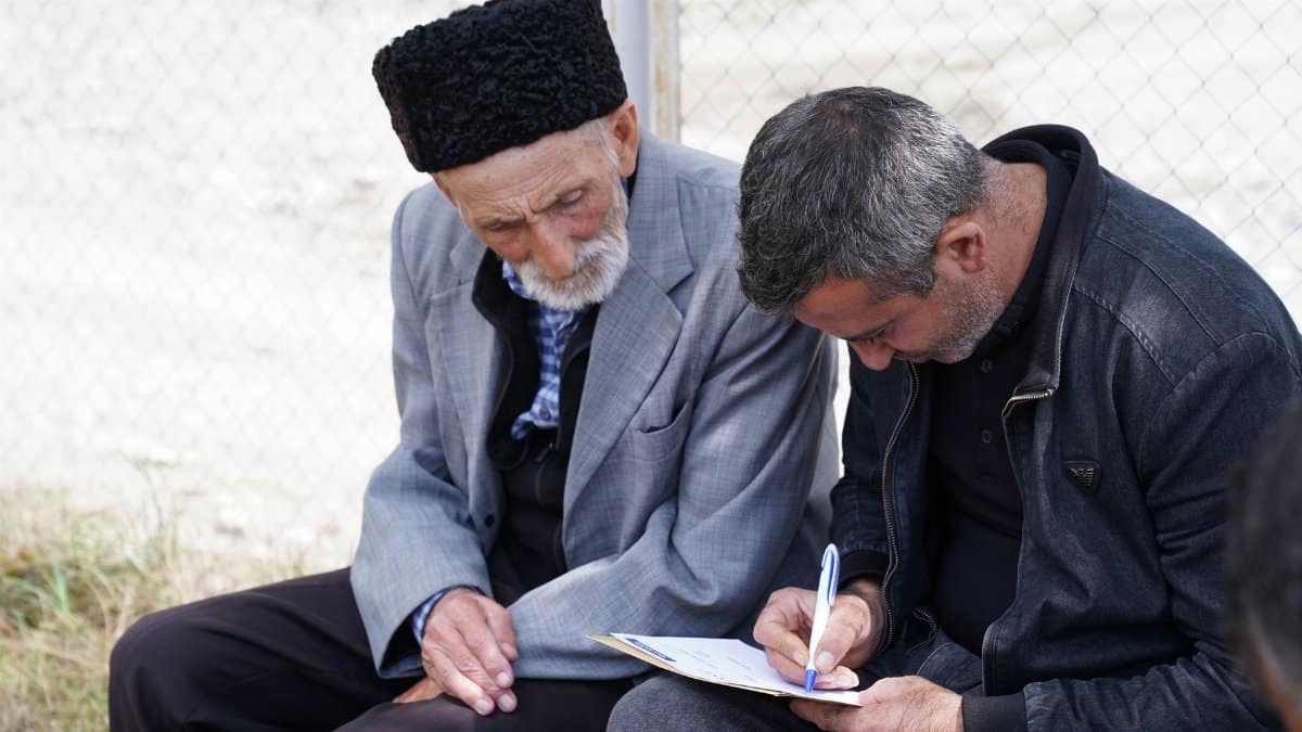 A man helps an elderly person fill out a form while sitting outdoors.