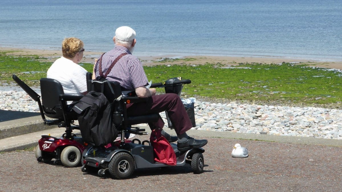 An elderly couple enjoys a seaside view while seated on mobility scooters, emphasizing leisure and accessibility.