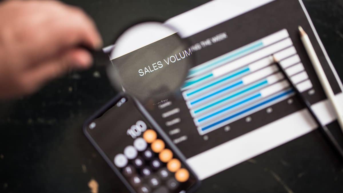 Close-up of a magnifying glass focusing on a sales volume chart next to a smartphone calculator app.