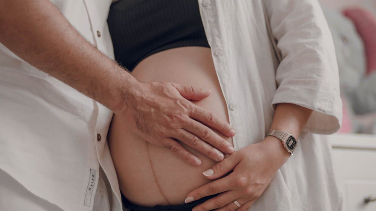Close-up of a pregnant couple in white shirts lovingly touching a baby bump indoors.