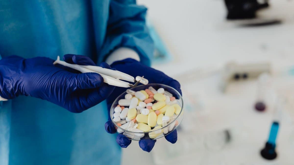 Scientist in gloves using tweezers to handle assorted pills in a petri dish.