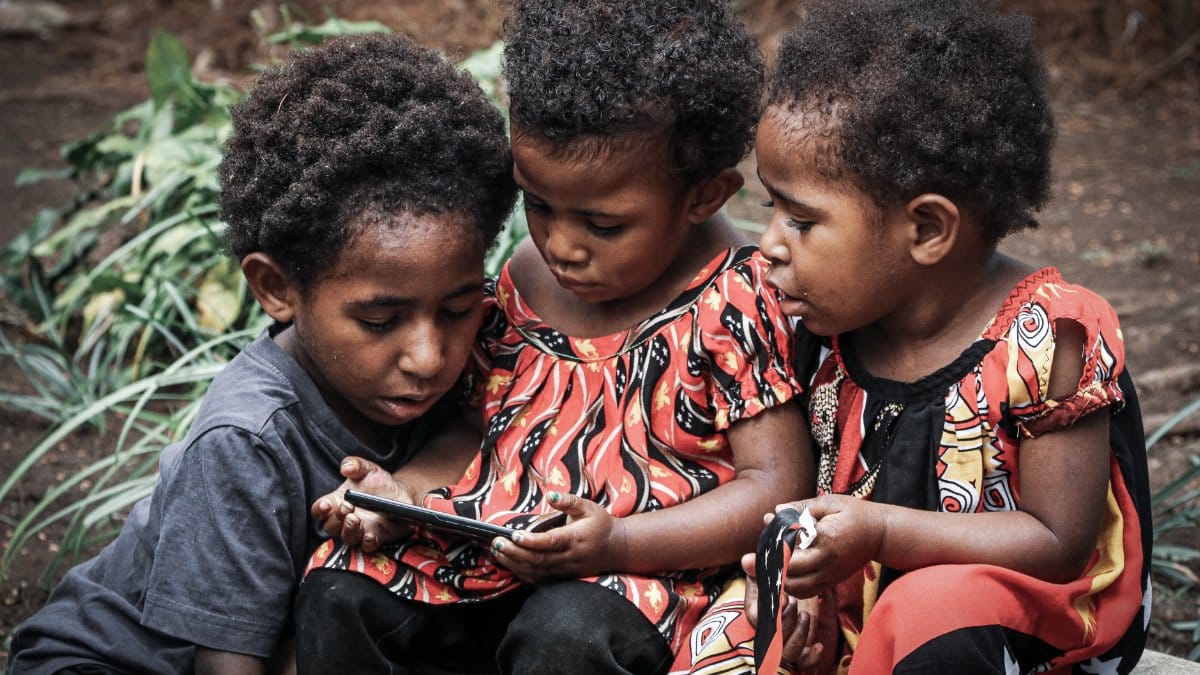 Three children using a smartphone outdoors in Papua New Guinea, exploring technology together.