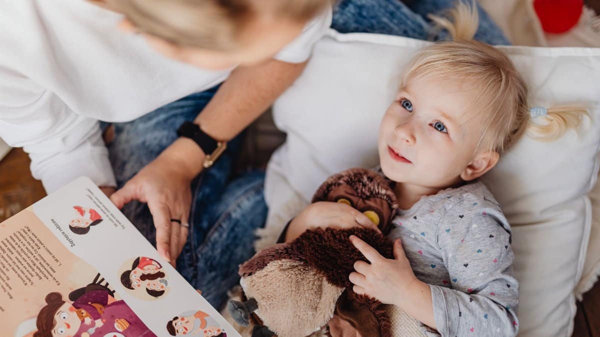 A mother reading a storybook to her toddler daughter indoors, depicting a cozy storytelling moment.