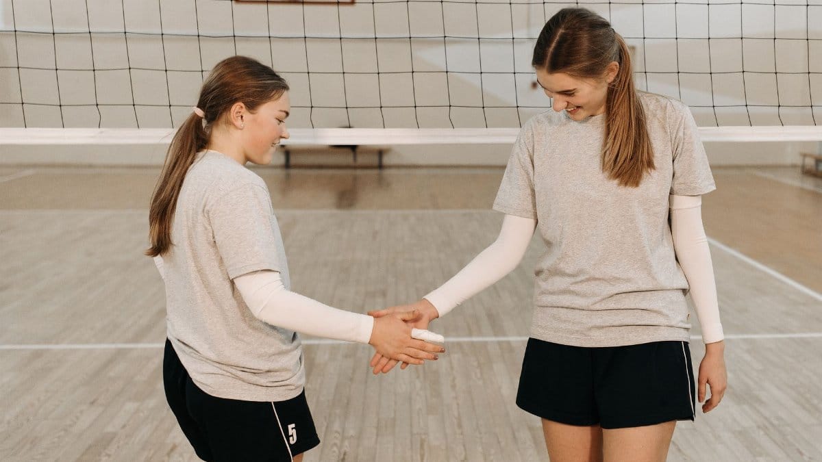 Two female volleyball players in sportswear shaking hands over the net inside a gym.