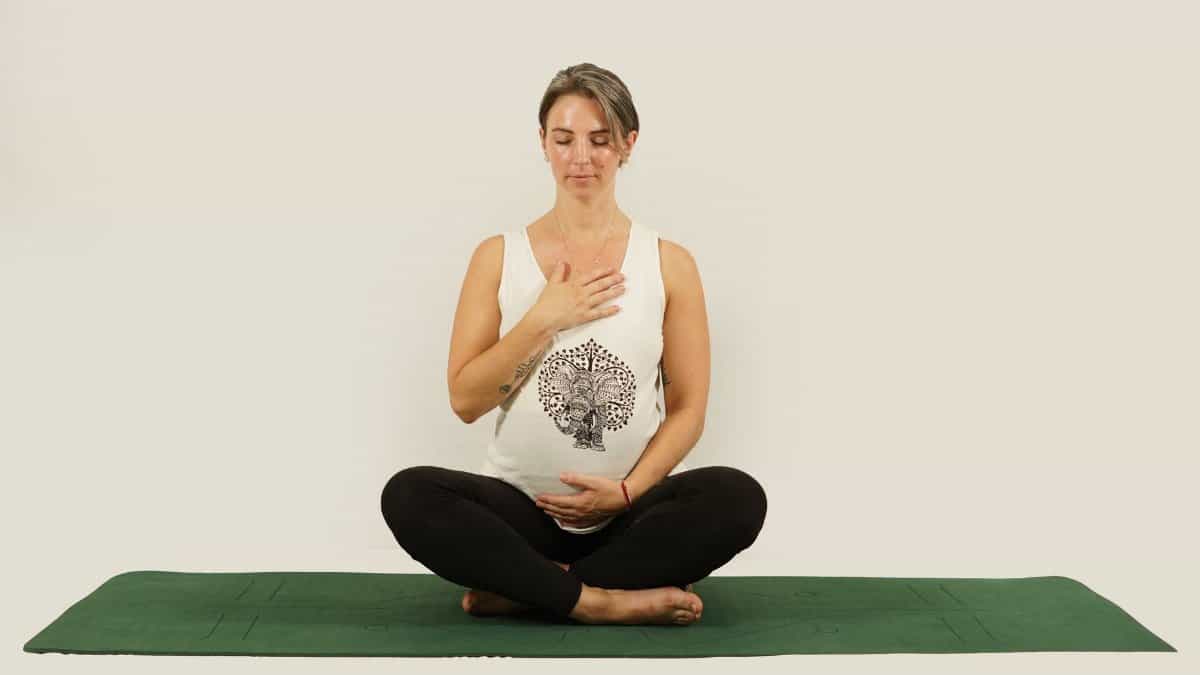 Pregnant woman sitting cross-legged on a green yoga mat, meditating with a calm expression indoors.