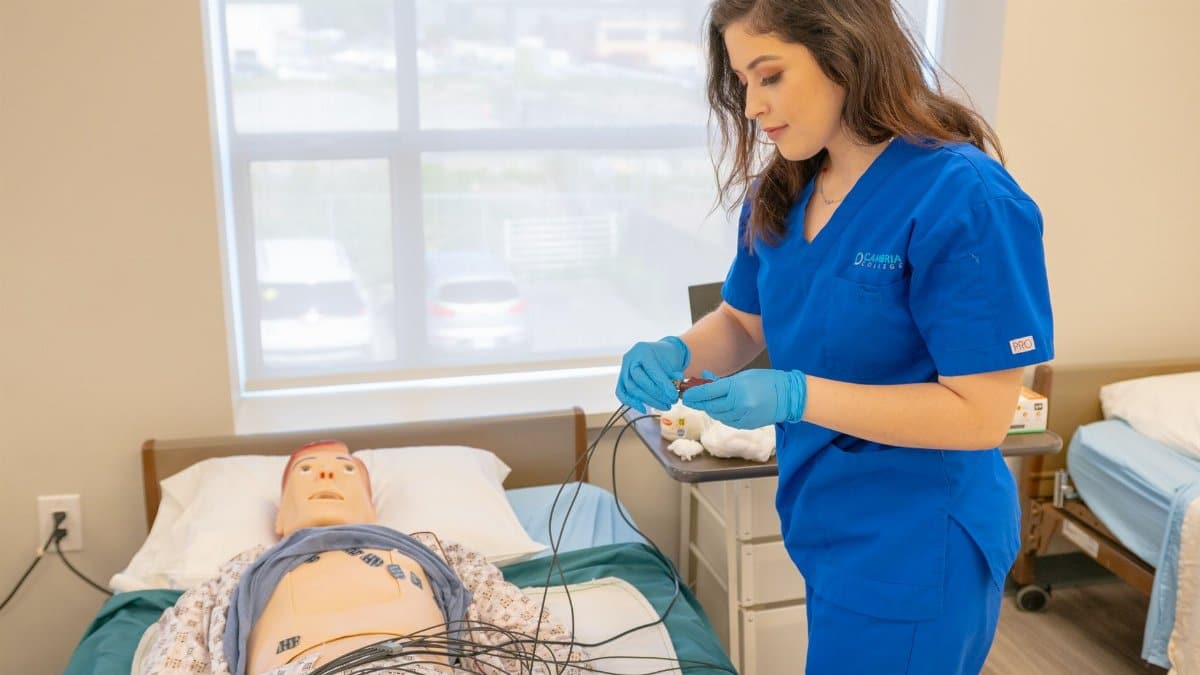 Medical student in blue scrubs practicing procedures on a patient training manikin in a clinical setting.