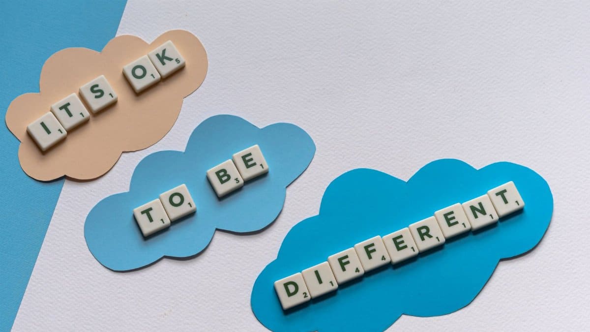 Scrabble tiles spell 'It's ok to be different' on colorful cloud cutouts, promoting diversity.
