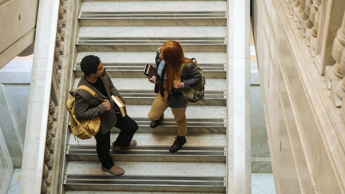Two students with backpacks and books talking on a marble staircase indoors.