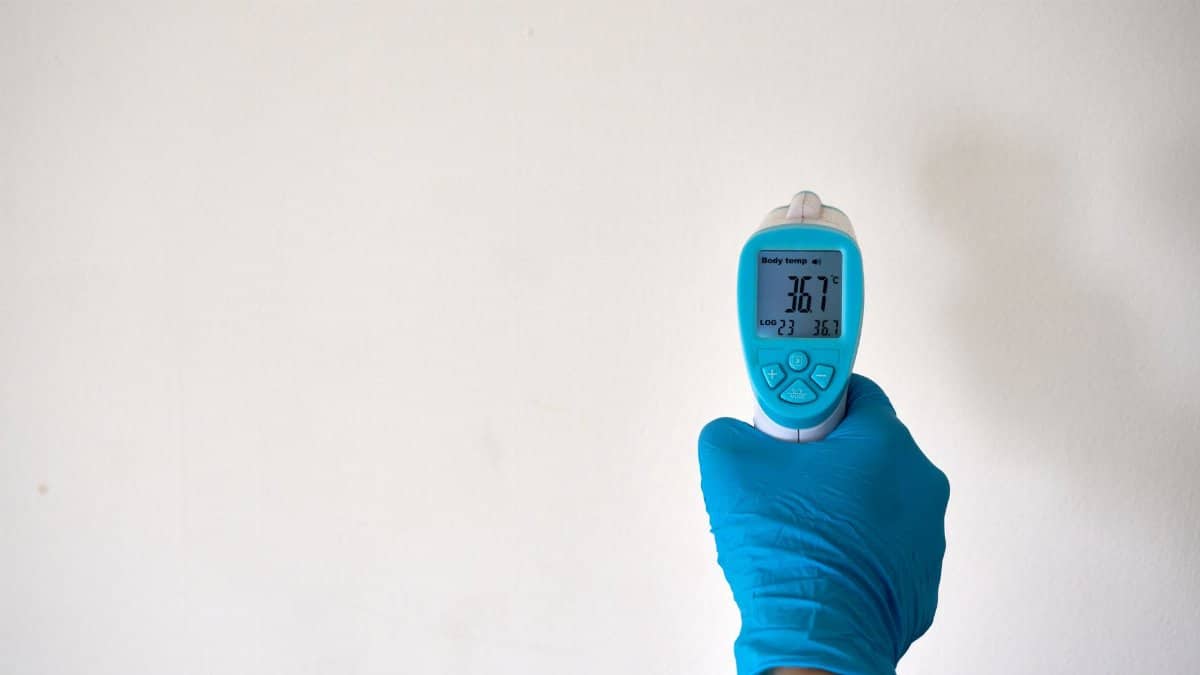 A hand in blue gloves holding an infrared thermometer against a plain background, signifying safety and health measures.