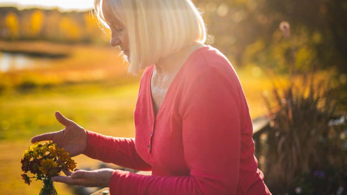 Elderly woman in garden admiring sunlit flowers, creating a peaceful autumn mood.