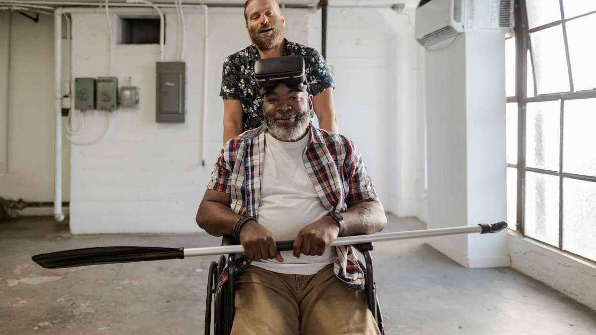 Two men enjoy virtual reality therapy indoors, one in a wheelchair holding a paddle.