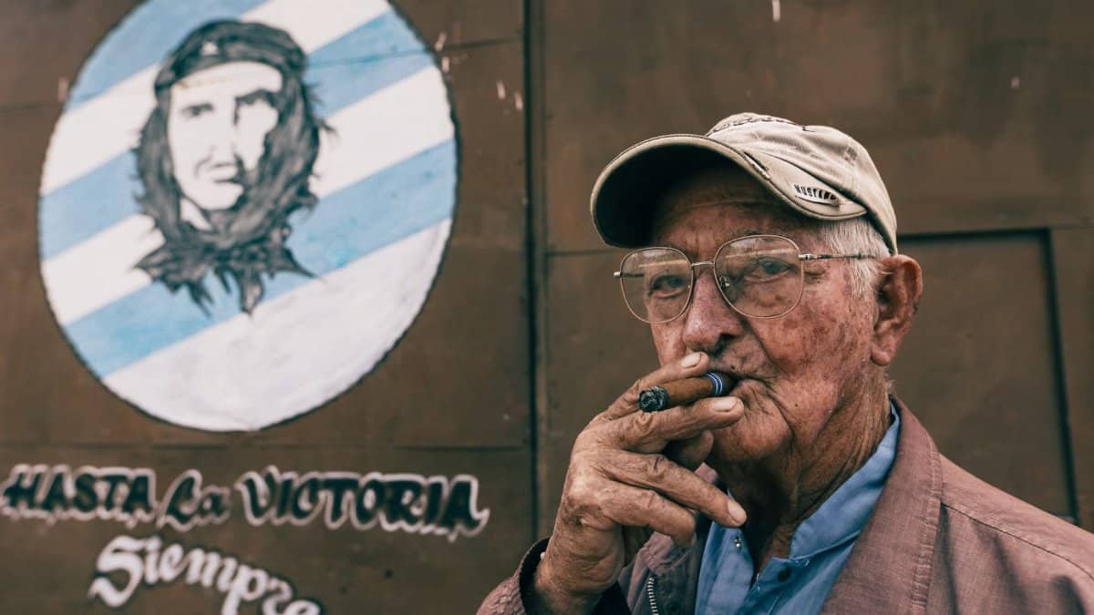Elderly man with eyeglasses smokes a cigar outdoors, standing by a Che Guevara mural.