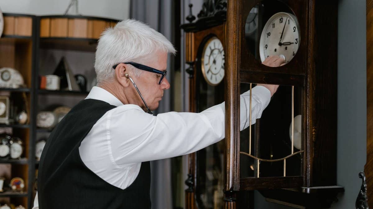 Elderly watchmaker in white shirt and vest tuning an elegant vintage clock indoors.