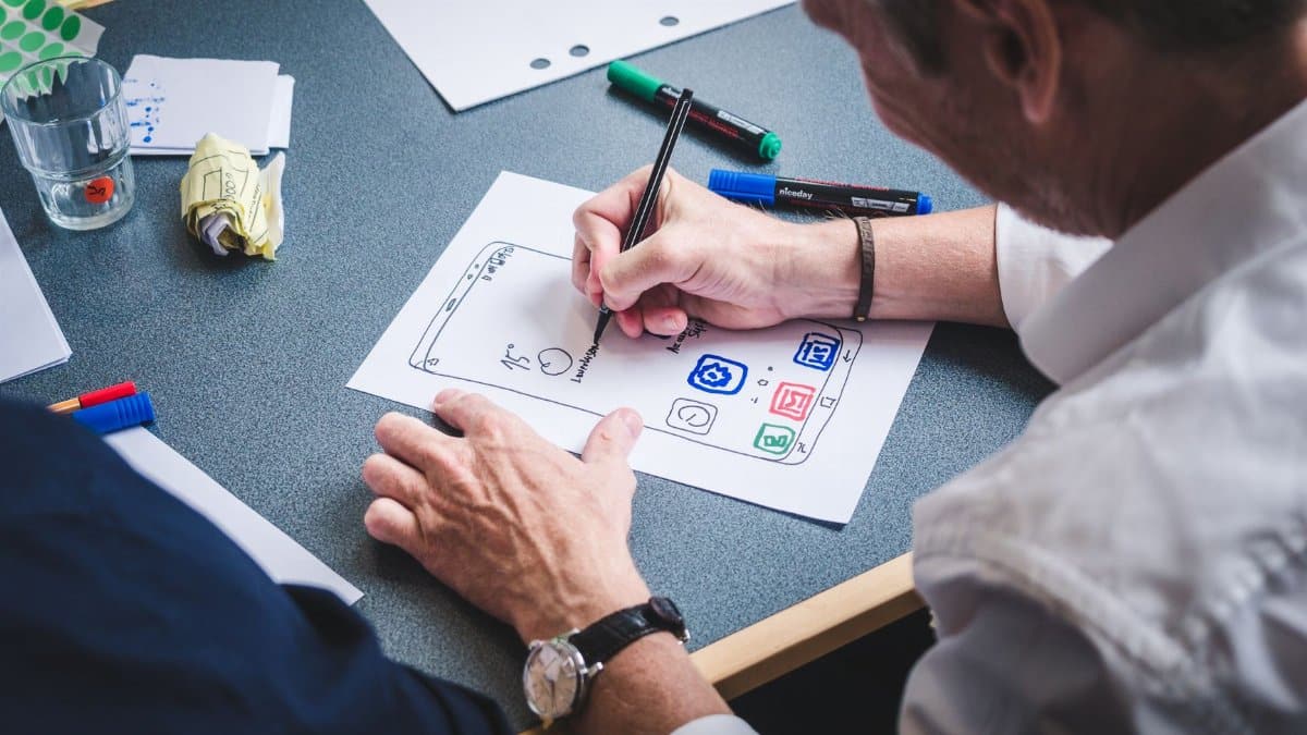 A designer sketches a mobile app prototype on paper at an office desk, focusing on creativity and design.