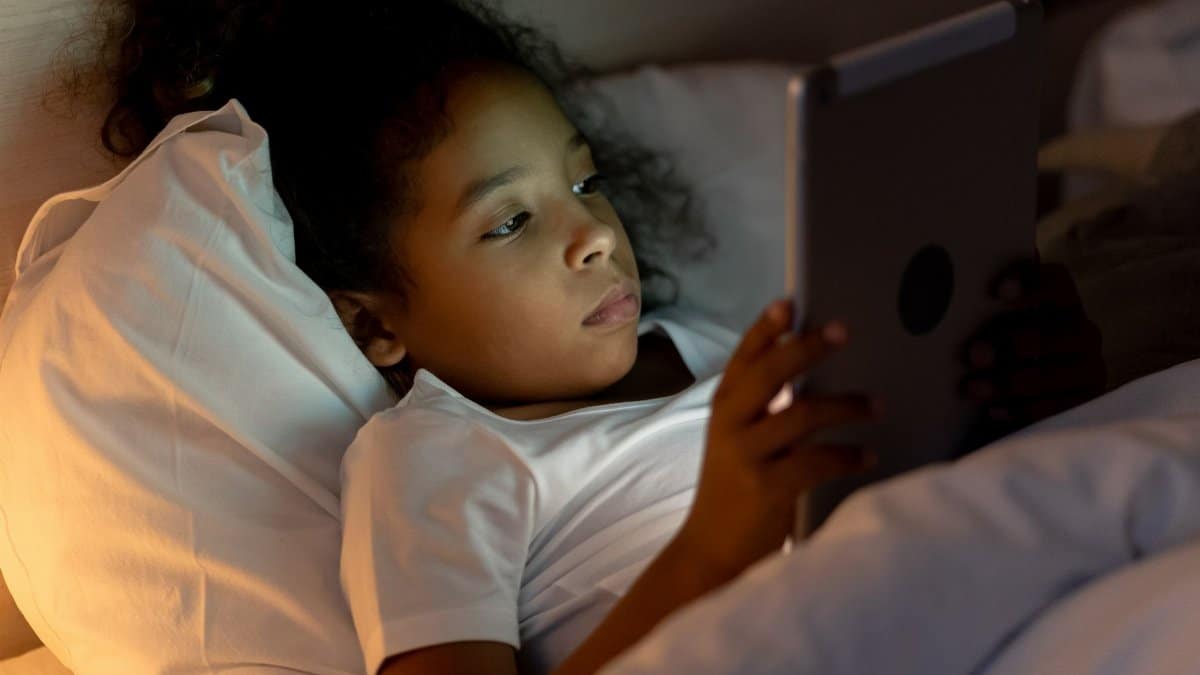 A young girl lying in bed, engrossed in a tablet screen at night, enjoying her bedtime routine.