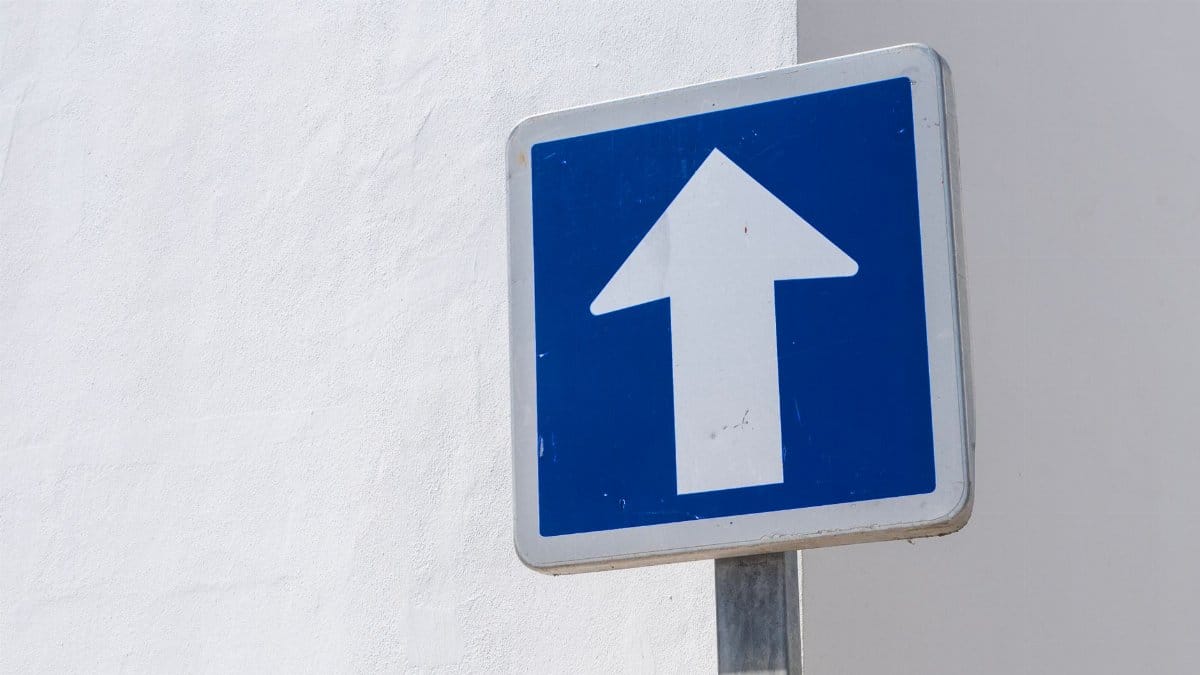 A simple blue directional traffic sign with white arrow over a white wall background, symbolizing direction or movement.