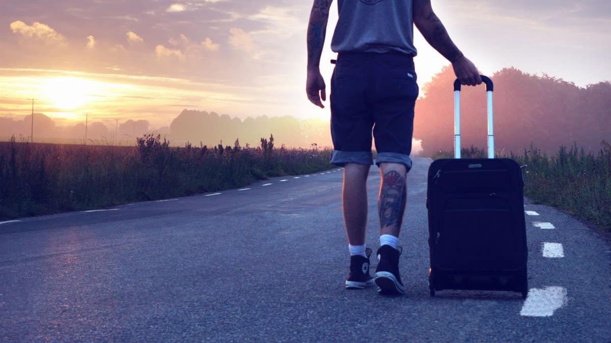 Man with tattoo walks along a rural road at sunrise with a suitcase, showcasing travel and adventure.