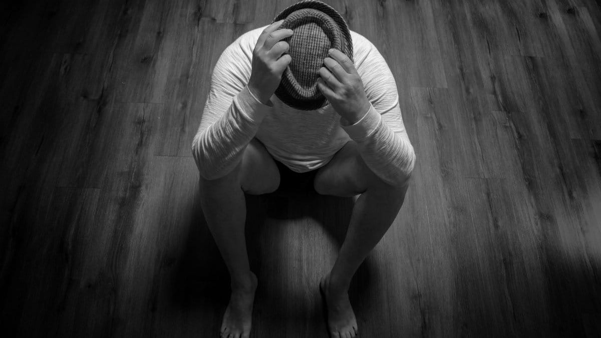 Black and white photo of a person sitting with a hat on a wooden floor, conveying sadness.