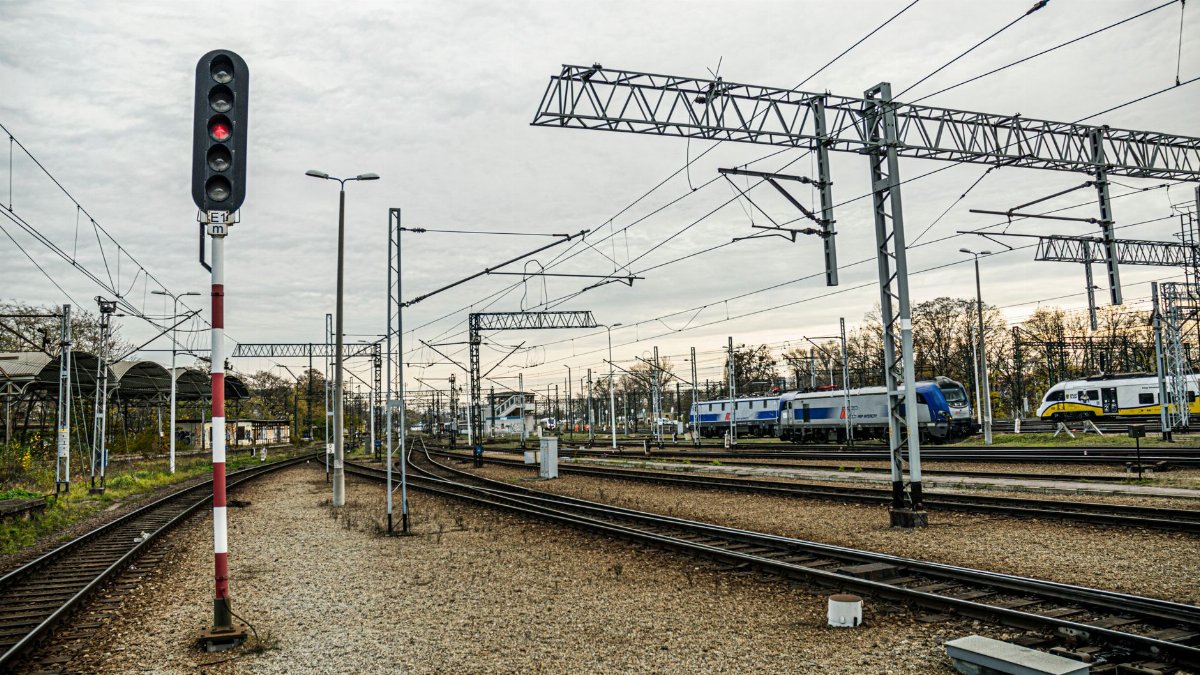 A modern train station with multiple tracks, trains, and a prominent signal under a cloudy sky.