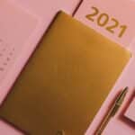A minimalist desktop with a 2021 calendar, golden notebook, and stationary on a pink background.