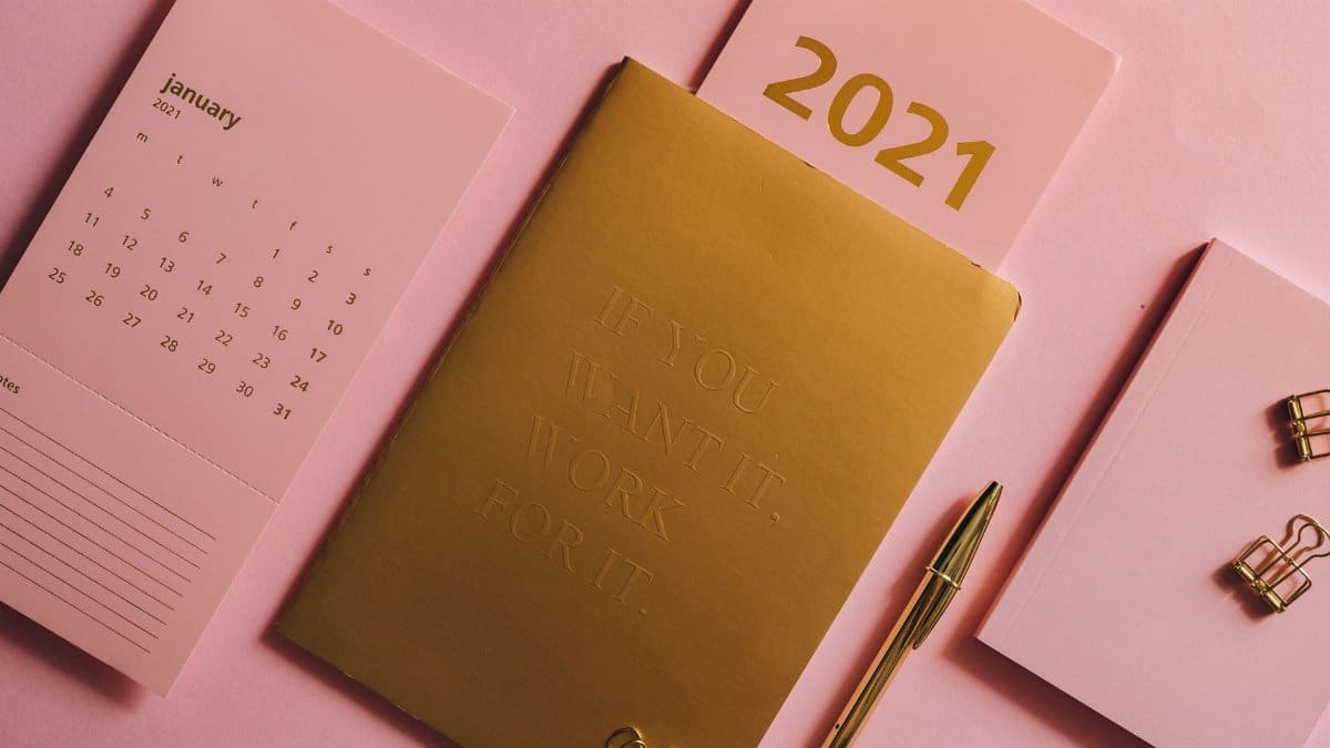 A minimalist desktop with a 2021 calendar, golden notebook, and stationary on a pink background.