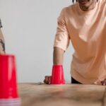 Two young men engaging in a fun cup game with red cups on a wooden table indoors.