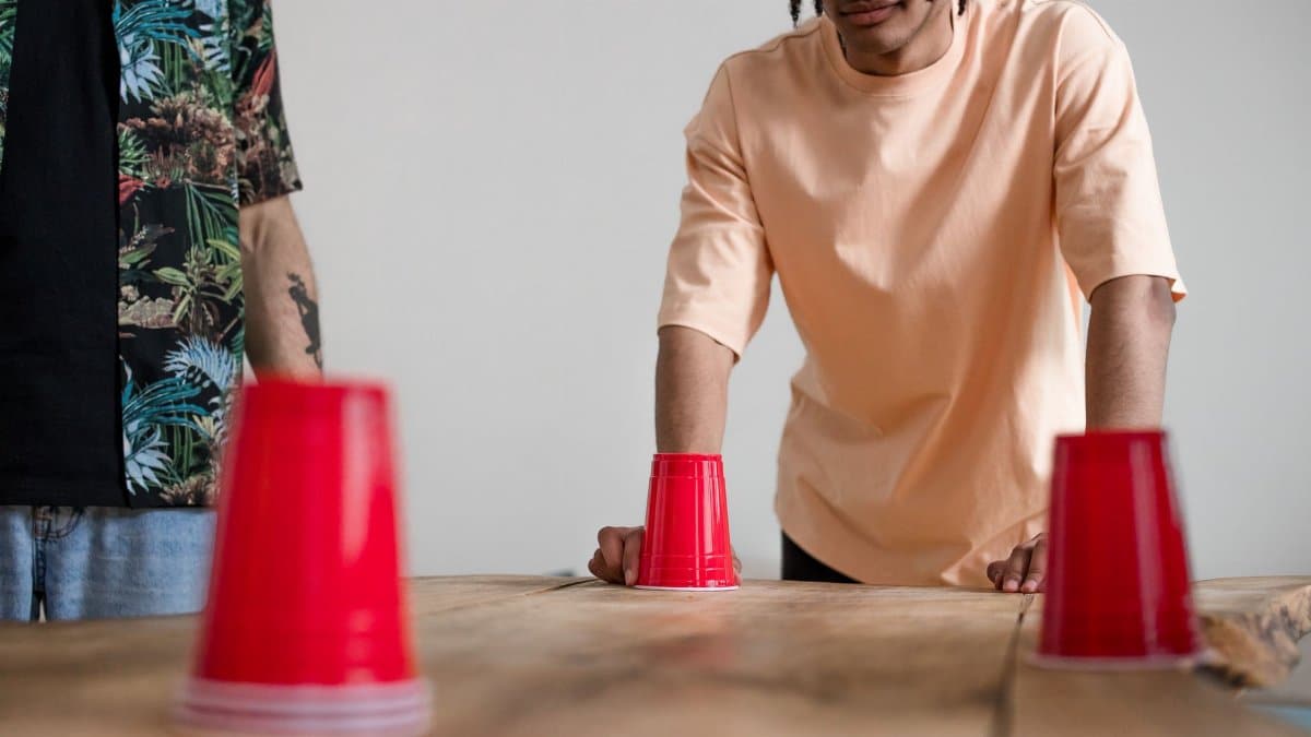 Two young men engaging in a fun cup game with red cups on a wooden table indoors.
