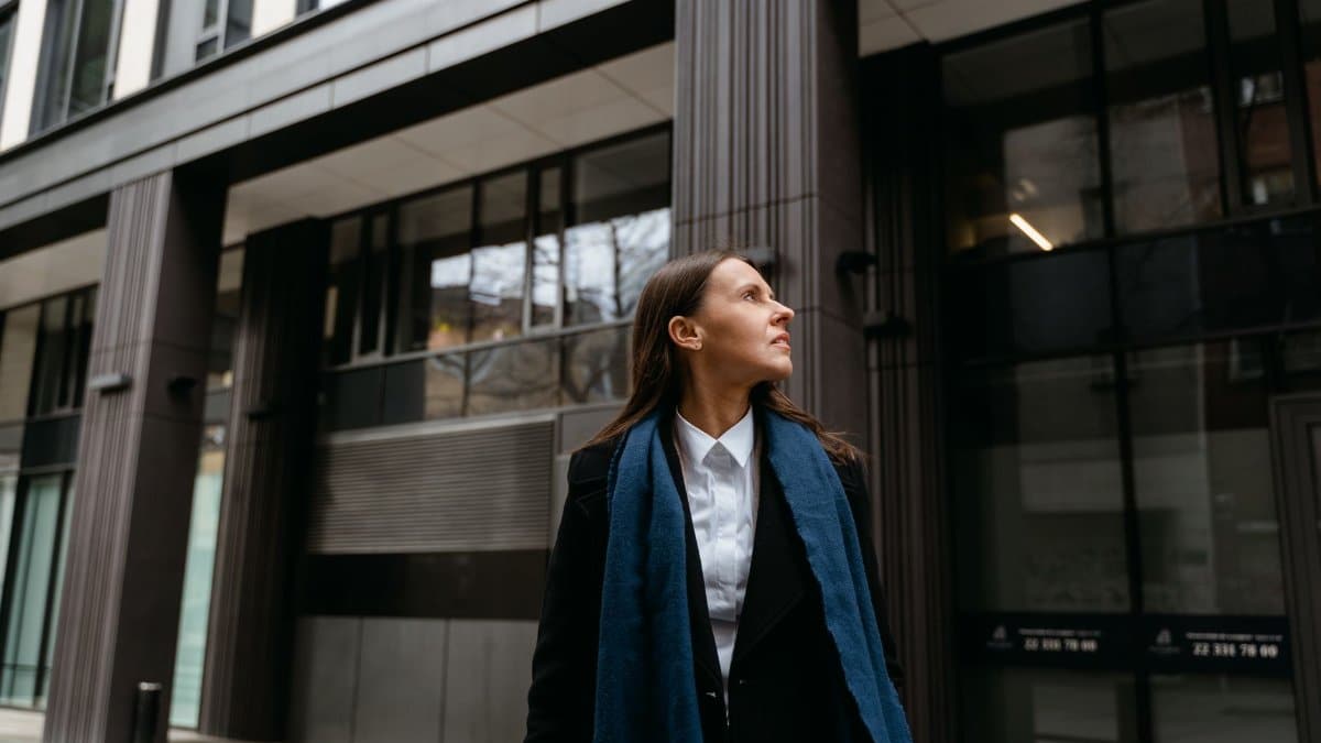 Woman in a black coat and blue scarf, looking up at a modern building facade.