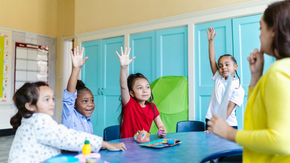 Children eagerly raising hands in a colorful classroom, engaged in learning.