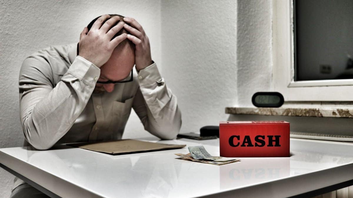 Bald man holding head in despair at desk with cashbox and money. Indoor office stress concept.