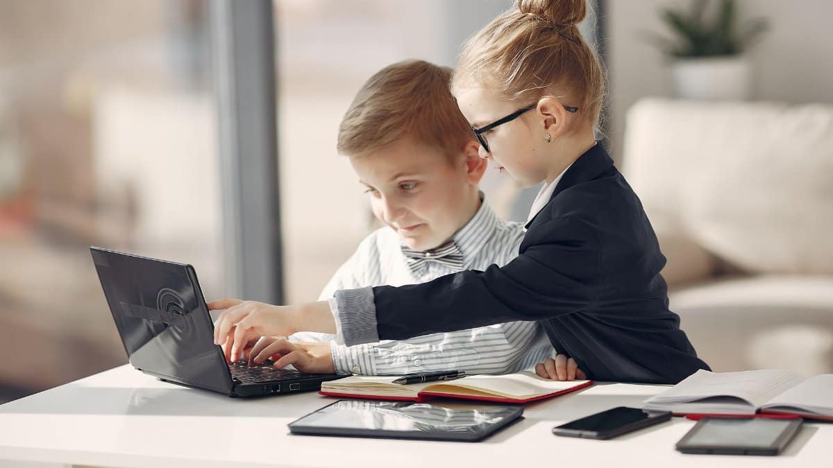 Side view of serious little girl pointing on laptop screen and explaining problem to coworker boy typing on keyboard in spacious room