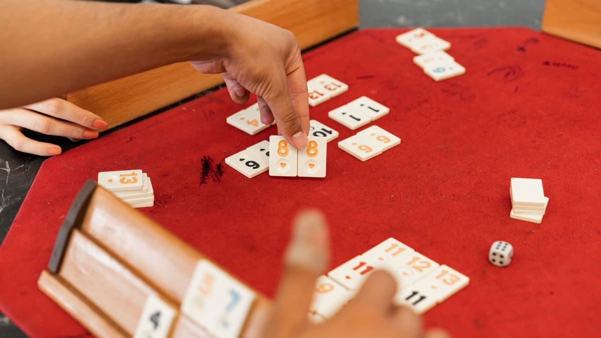 Close-up of hands playing Rummikub with numbered tiles on a red felt table.