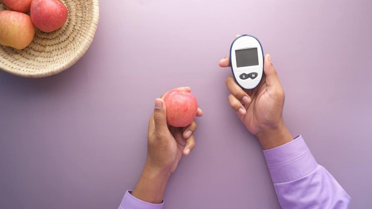 Hands holding apple and glucometer on purple surface, symbolizing health balance.