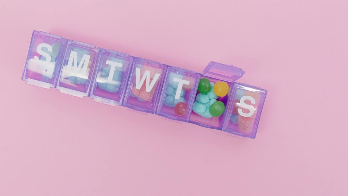 Vibrant candies arranged in a weekly pill organizer on a pastel pink background.